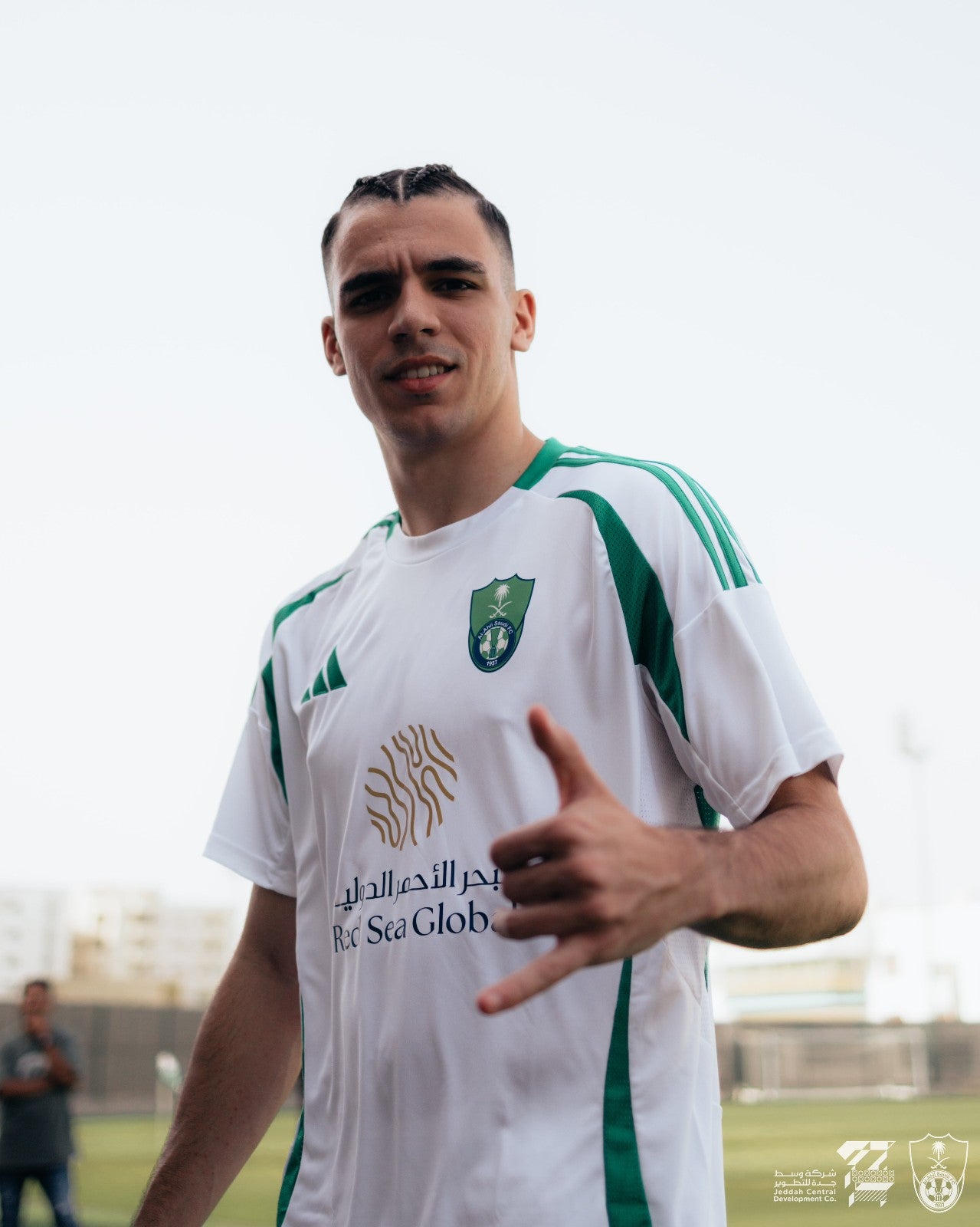 HAFLxfxaxIoTomy | UAE SPORT STORE Smiling young man in a white jersey with green stripes giving a thumbs up on a sports field, crest on chest visible.