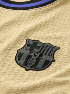 Close-up of a black and gray FC Barcelona shield patch sewn onto beige textured jersey fabric.
