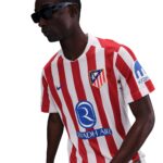 Man wearing a red-and-white striped soccer jersey with club crest on the chest and sponsor logos, facing left while wearing sunglasses.