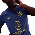 Person wearing a blue Riyadh Air football jersey with a gold sponsor logo and crest, posed with hand by head.