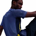 Man wearing a blue athletic shirt stretches his arm to the side, viewed from the back/side profile