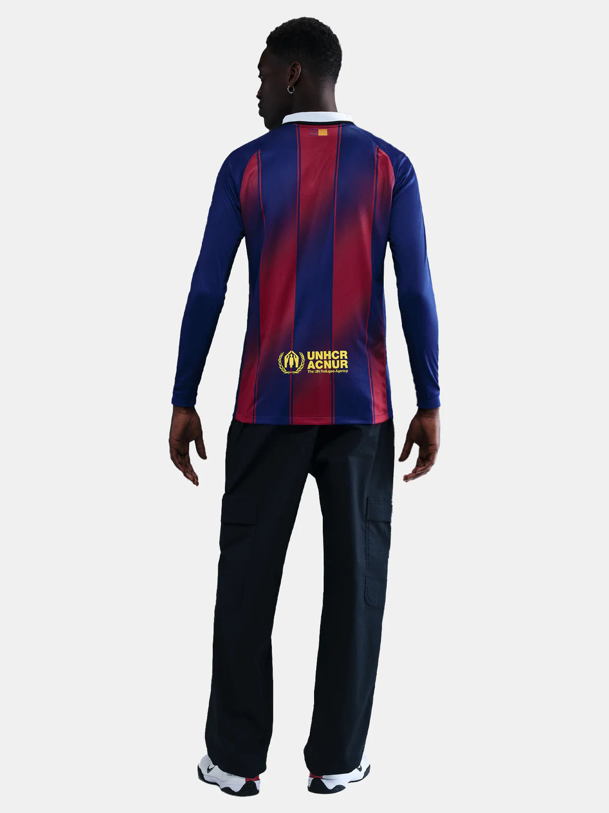 HJ4611-456-PHSBMG01_copia | UAE SPORT STORE Back view of a man in a blue and red striped long-sleeve shirt with UNHCR logo on the lower back, wearing black cargo pants and white sneakers.