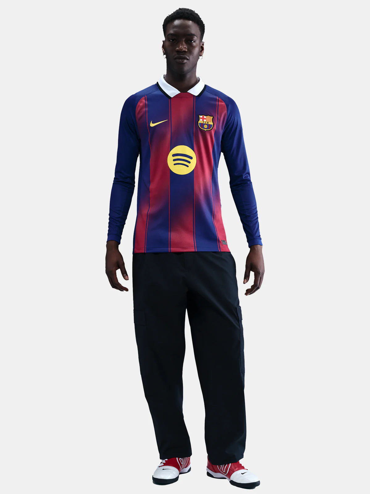 HJ4611-456-PHSFMG01_copia | UAE SPORT STORE Full-body shot of a man wearing a blue and red FC Barcelona jersey with a yellow sponsor logo, black pants, and red-and-white sneakers against a light background.