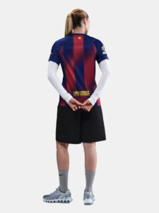 Back view of a person wearing a blue-red striped soccer jersey with UNHCR logo on lower back and a small flag on the neck area, black shorts, gray socks, and light-blue cleats.