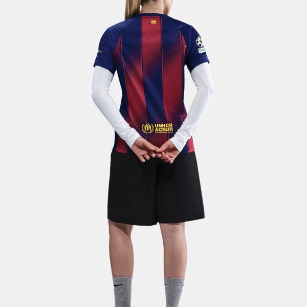 Back view of a person wearing a blue-red striped soccer jersey with UNHCR logo on lower back and a small flag on the neck area, black shorts, gray socks, and light-blue cleats.