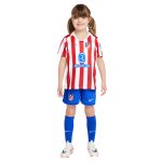 Young girl in an Atlético Madrid soccer kit: red-and-white striped jersey with blue shorts and blue socks, smiling at the camera.