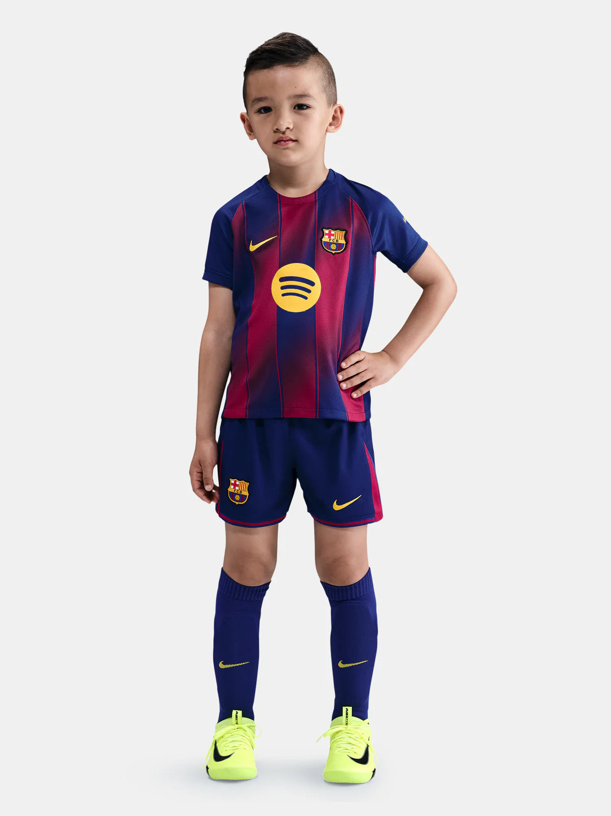 HJ5619-456_415095256_D_A_1X1_c2197d38-dfbd-4907-abd7-98264fb5092d Young boy models a FC Barcelona youth soccer kit with blue‑red striped shirt, shorts, and neon yellow cleats.