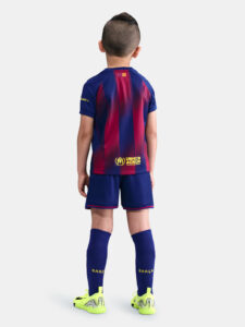 Boy wearing a Barcelona football kit seen from the back, blue and red striped jersey with UNHCR logo and blue shorts.