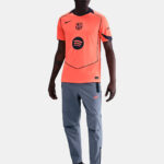 Soccer player modeling a bright orange training jersey with a Barcelona crest, sponsor logo, dark piping, gray pants, and black sneakers.