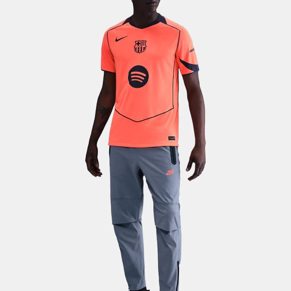 Soccer player modeling a bright orange training jersey with a Barcelona crest, sponsor logo, dark piping, gray pants, and black sneakers.