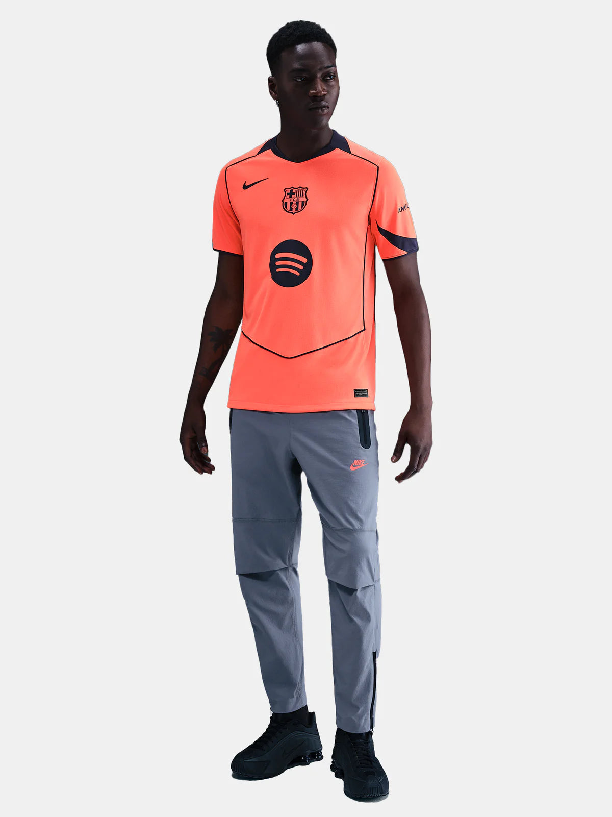HM3201-855_415229025_D_D_1X1_dcfab875-933d-4a8e-85e5-ff5c817297b5 Soccer player modeling a bright orange training jersey with a Barcelona crest, sponsor logo, dark piping, gray pants, and black sneakers.