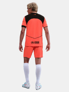 Person seen from behind in a bright orange soccer jersey and shorts with a black upper back panel; UNHCR logo on the lower back.