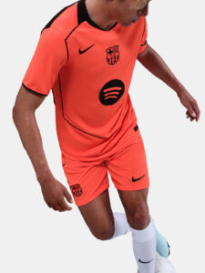 Soccer player in neon orange FC Barcelona jersey and shorts, with Barca crest, Nike swoosh, and Spotify logo, dribbling a white ball.