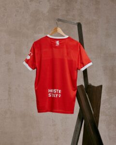 Red sports jersey hanging on a wooden hanger mounted on a metal rack against a textured wall, showing white cuff trim and logos on back neck and lower back area.