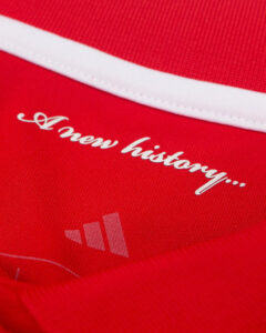 Close-up of red fabric with white cursive text 'A new history...' and a small light Adidas logo.