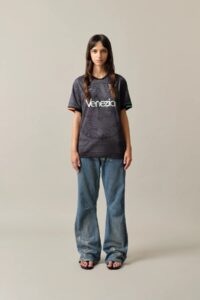 HomeW_Front1_299c1d40-1916-44a0-9297-1ffce4338dcf | UAE SPORT STORE شابة تقف في استوديو محايد ترتدي تيشيرت فينيتسيا باللون الأسود-الرمادي وجينز واسعًا وصندلًا، وتواجه الكاميرا.