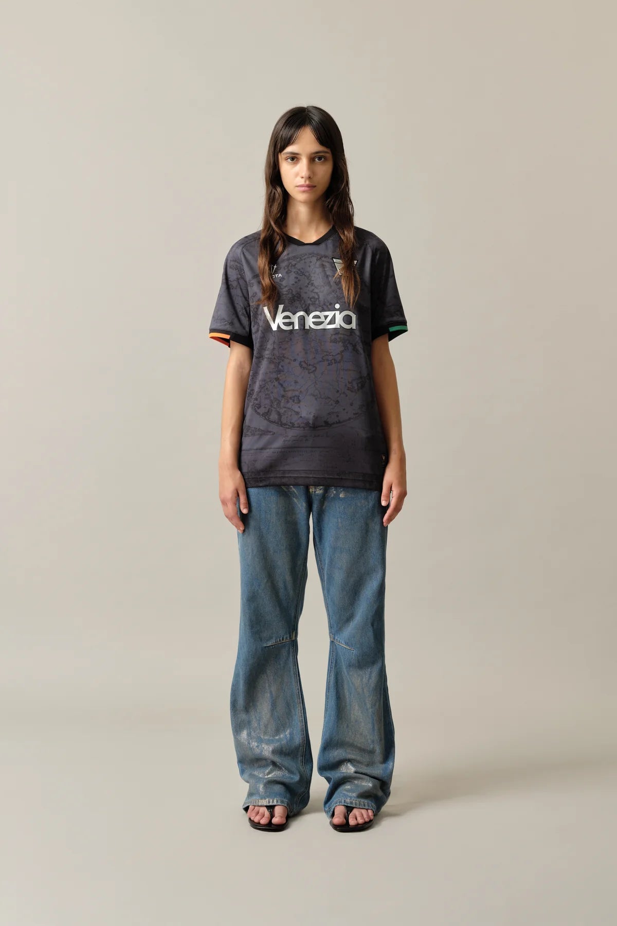 HomeW_Front1_299c1d40-1916-44a0-9297-1ffce4338dcf | UAE SPORT STORE شابة تقف في استوديو محايد ترتدي تيشيرت فينيتسيا باللون الأسود-الرمادي وجينز واسعًا وصندلًا، وتواجه الكاميرا.
