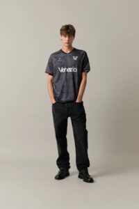 Home_Front1_bad00eba-9fba-45e2-a852-a39913c53e9b | UAE SPORT STORE شاب يرتدي قميص كرة قدم فينيتسيا باللون الرمادي الداكن مع جينز أسود، واقفًا ويداه في الجيوب أمام خلفية محايدة.