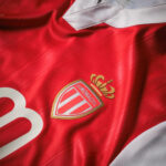Close-up of a red AS Monaco soccer jersey with a white collar and the club crest in gold and silver.
