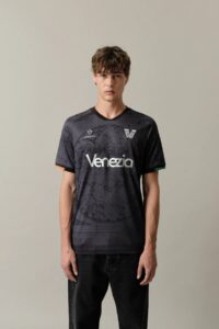 Home_Jersey_Venezia_3_fb70cb3e-e68f-465b-899a-8f4d74ae6e83 | UAE SPORT STORE شاب ذو شعر مموّج يرتدي قميص كرة قدم فينيتسيا باللون الرمادي الداكن، واقفًا أمام خلفية فاتحة بسيطة.