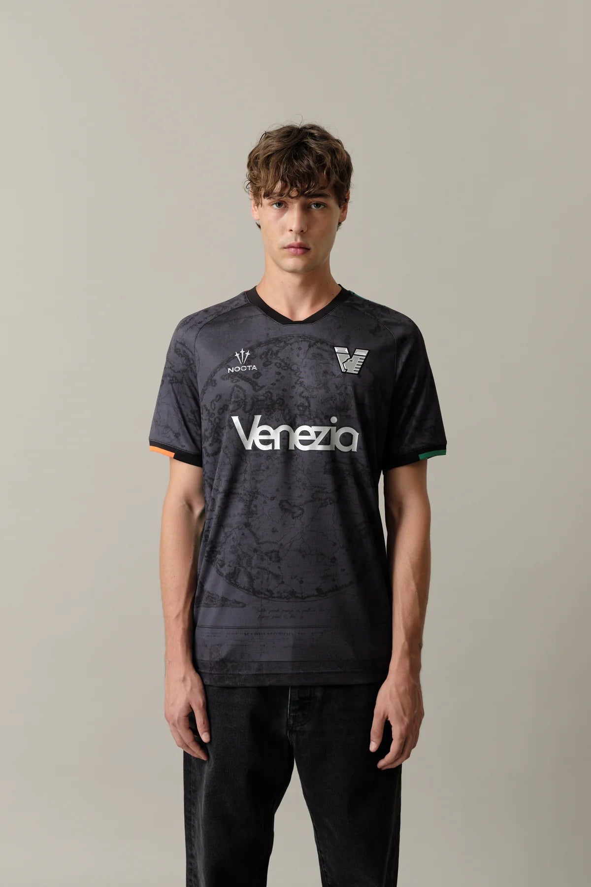 Home_Jersey_Venezia_3_fb70cb3e-e68f-465b-899a-8f4d74ae6e83 | UAE SPORT STORE شاب ذو شعر مموّج يرتدي قميص كرة قدم فينيتسيا باللون الرمادي الداكن، واقفًا أمام خلفية فاتحة بسيطة.