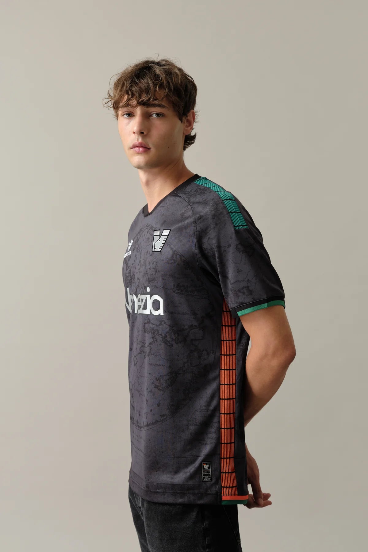 Home_Side_ad0bb08d-964a-4174-a2b4-83019a7a1d3d | UAE SPORT STORE عارض رجل يرتدي قميصًا رياضيًا باللون الرمادي الداكن مع شريط جانبي برتقالي ولمسات خضراء، يظهر من الجانب أمام خلفية محايدة.