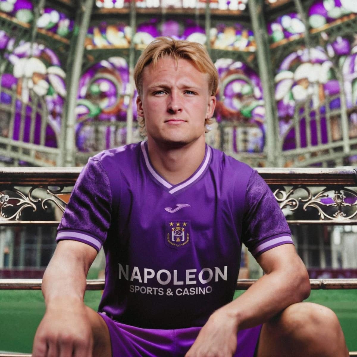 Young man in a purple Napoli sports jersey sits on a bench with a colorful patterned background behind him (stadium or venue).
