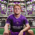 Young man in a purple Napoli sports jersey sits on a bench with a colorful patterned background behind him (stadium or venue).