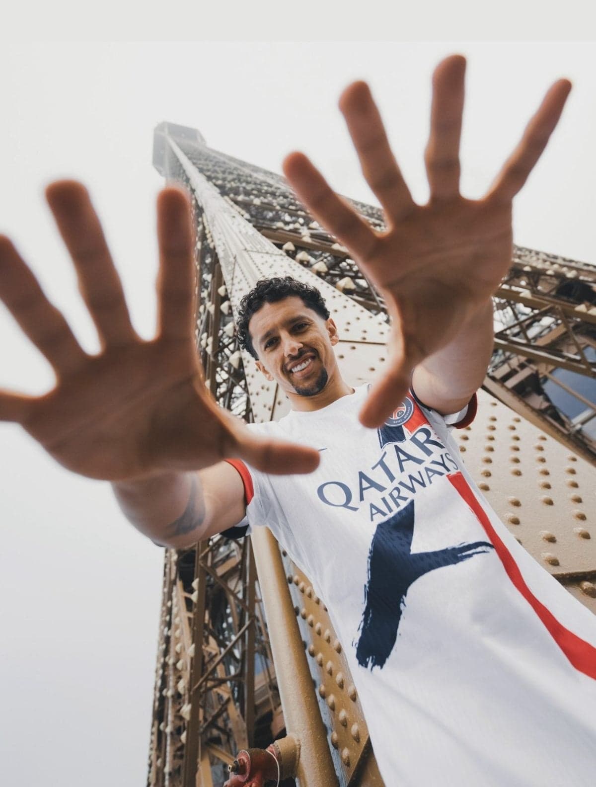 Man in a Qatar Airways shirt smiles as he poses for a selfie, hands reaching toward the camera from a tall riveted steel bridge structure.