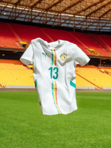 White soccer jersey with number 13 and colorful vertical embroidery, crest on chest, held up in a stadium with orange seats.