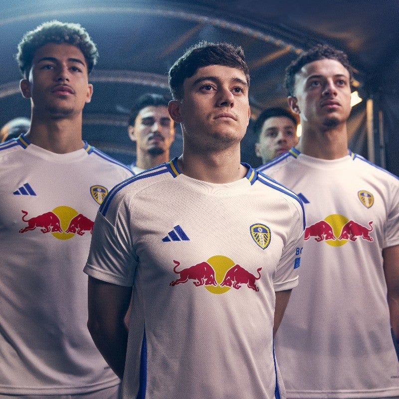 IDvTt7sqEZrwhmC | UAE SPORT STORE Three young soccer players in white jerseys with Red Bull logo stand in a formation, looking focused in a stadium corridor under blue lighting.