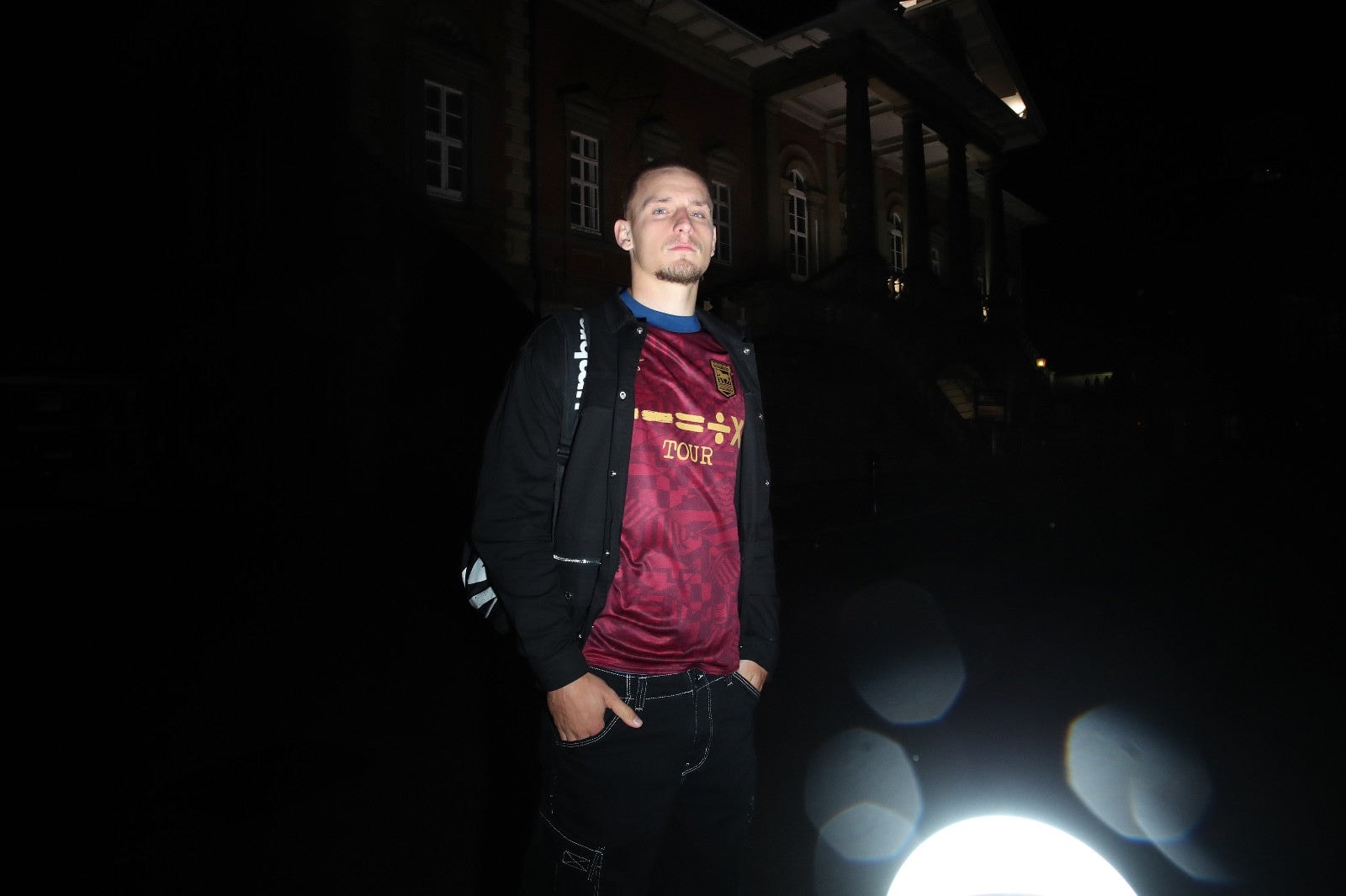 IM0P9o4uzGXxyrj | UAE SPORT STORE Man posing outside at night in front of a lit, columned building, wearing a maroon tour jersey and a backpack.
