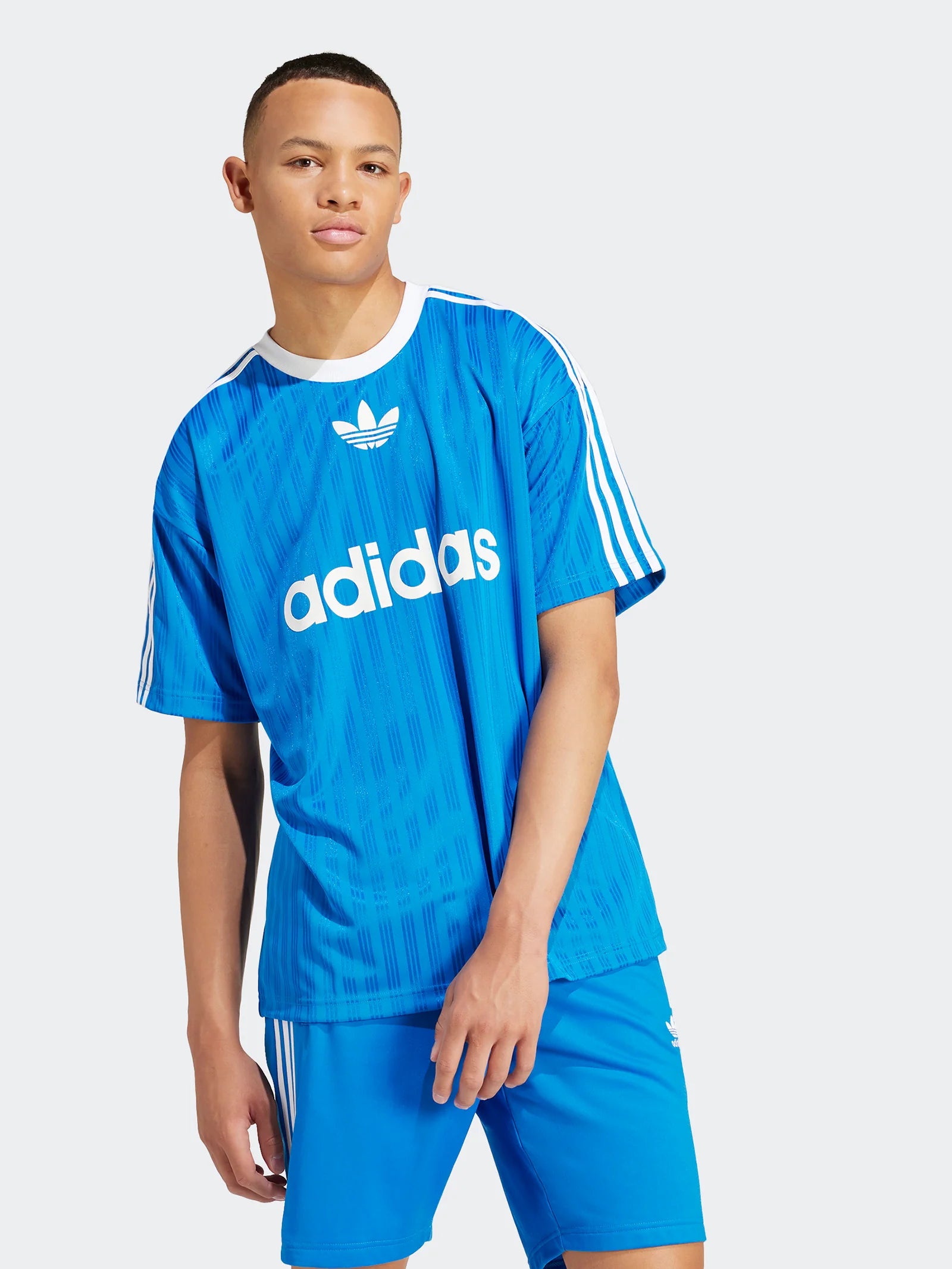 IM9456_6_APPAREL_OnModel_WalkingView_grey_2048x_4cf2ab69-e93a-4bc1-b989-afd7cb6b05e1 | UAE SPORT STORE Young man wearing a bright blue Adidas jersey with white stripes, Adidas logo, and bold 'adidas' text, standing against a light gray background.