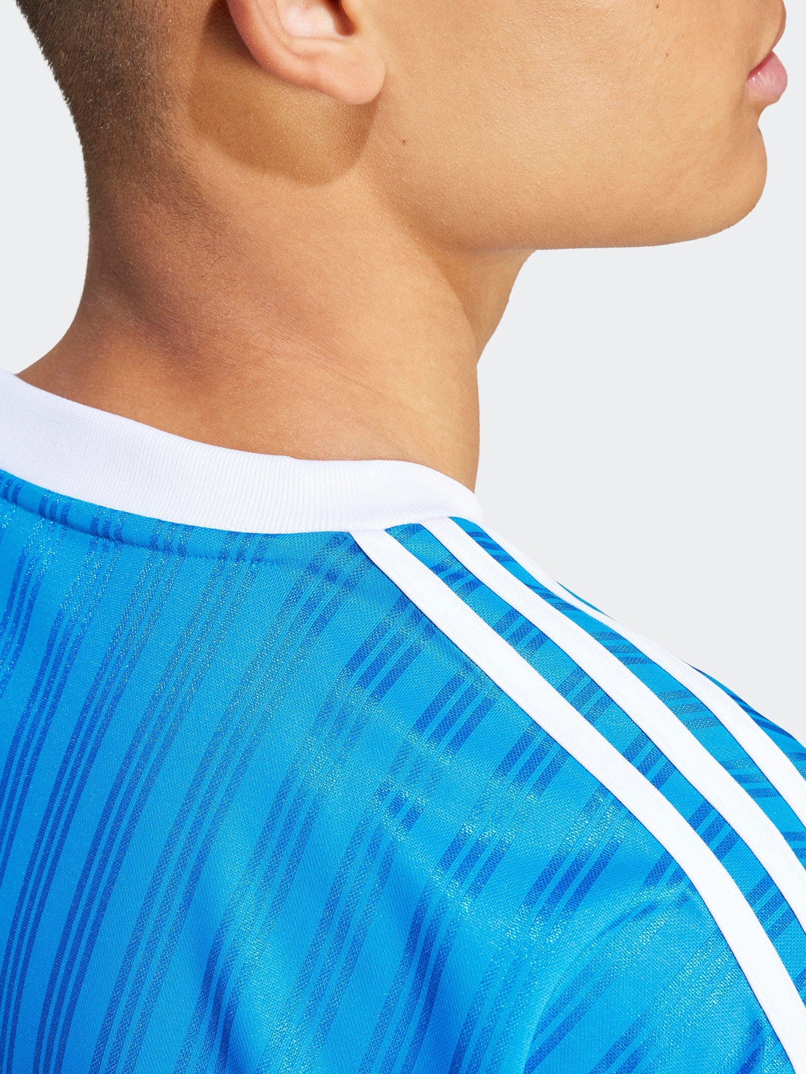 IM9456_8_APPAREL_OnModel_DetailView2_grey_2048x_9ae5dd8f-b513-44a3-88f0-ba420d674d6f | UAE SPORT STORE Close-up of a person’s neck and jawline wearing a blue striped athletic jersey with white trim.