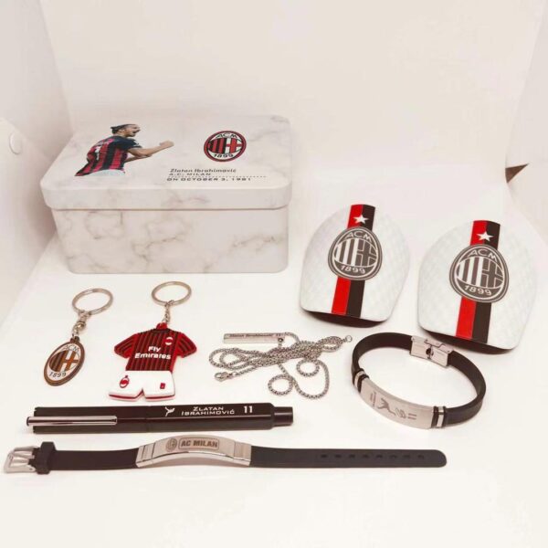 AC Milan memorabilia laid out on a white surface: tin box with Zlatan Ibrahimović image, two Milan crest cards, keychains, a red-and-black mini jersey, bracelet, necklace, pen and watch bands.