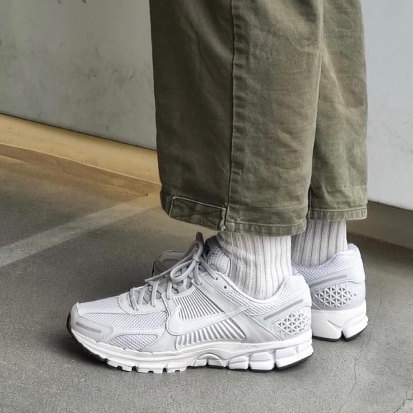 Close-up of white athletic sneakers with ribbed socks and olive pants, standing on a concrete floor.
