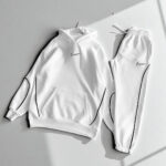 White Nike tracksuit: a hoodie with front pocket and drawstring hood paired with matching jogger pants featuring black piping accents.