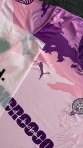 Close-up of a pink and purple athletic jersey with abstract camo shapes; a translucent plastic bag rests over the fabric with a circular emblem in the corner.