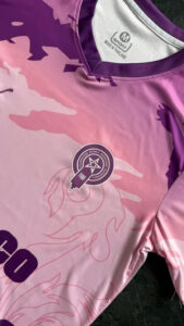 Pink and purple sports jersey with camo pattern and a circular embroidered badge featuring a star and ribbon motif on the chest area.