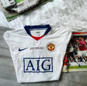 White Manchester United football jersey on a bed, featuring the AIG sponsor, club crest, and Nike swoosh.