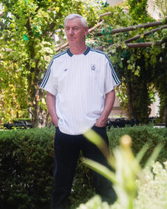 Older man with white hair standing outdoors in a garden, wearing a white pinstripe athletic shirt.