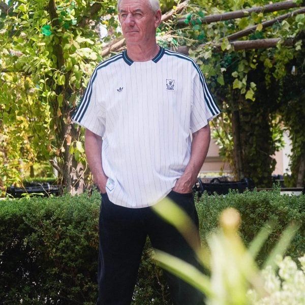 Older man with white hair standing outdoors in a garden, wearing a white pinstripe athletic shirt.