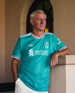 Older man wearing a turquoise Liverpool FC jersey leans on a beige railing indoors, looking to the right.