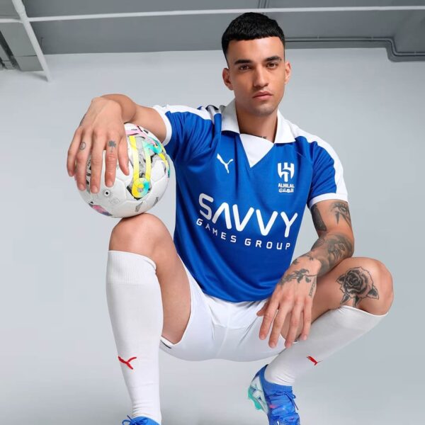 Soccer player in a blue and white kit crouches with a soccer ball resting on his knee, showcasing the Savvy Games Group logo on the chest.