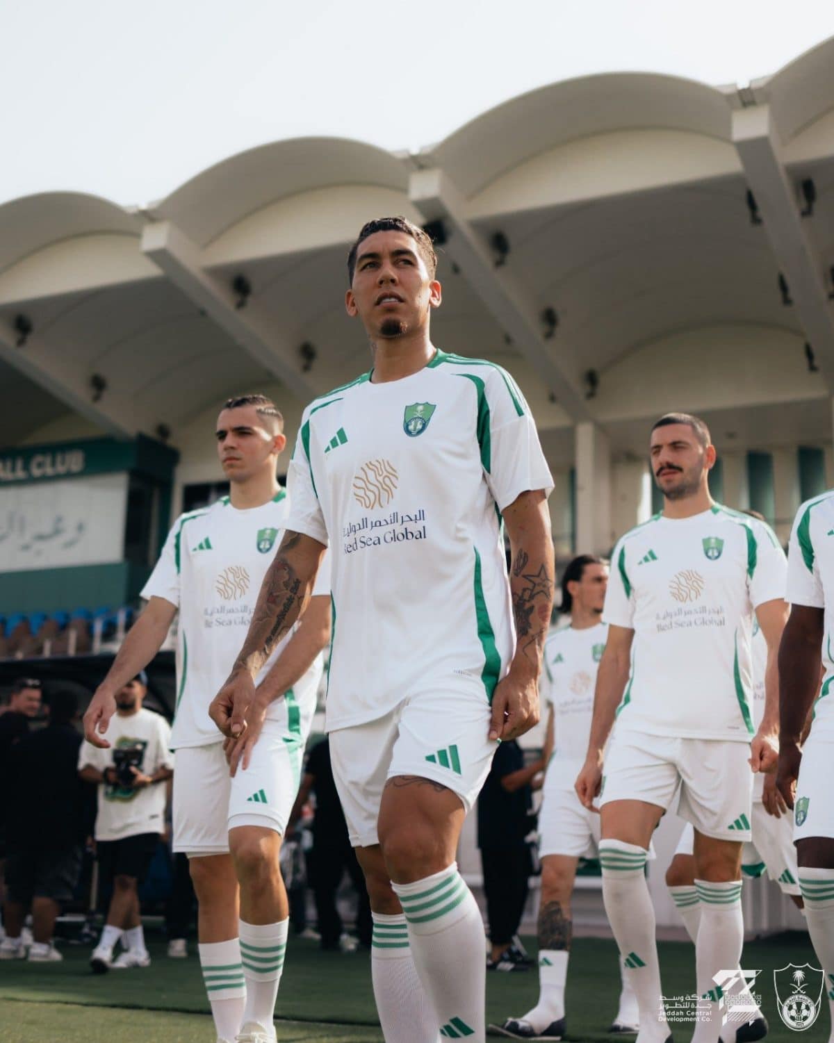 Soccer players in white and green uniforms walk onto the field, led by a central player, with a stadium backdrop behind them.