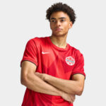 Portrait of a young man in a red Canada soccer jersey with arms crossed, looking confident against a light gray background.