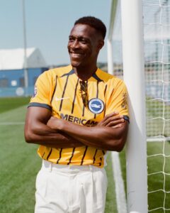 Smiling man in a yellow striped Brighton & Hove Albion jersey leaning against a soccer goal post on a sunny field, with sunglasses hanging from his jersey neckline.