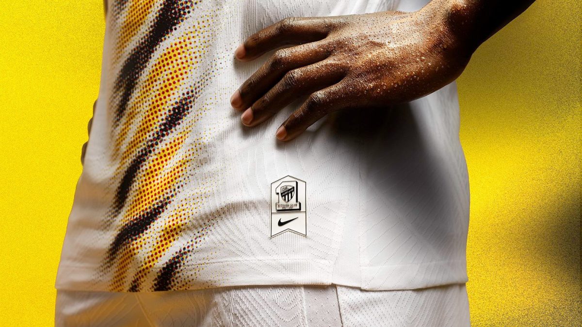 Close-up of a person in a white textured sports jersey with a yellow, black pattern, hand on hip, badge featuring a logo and Nike swoosh against a yellow background.