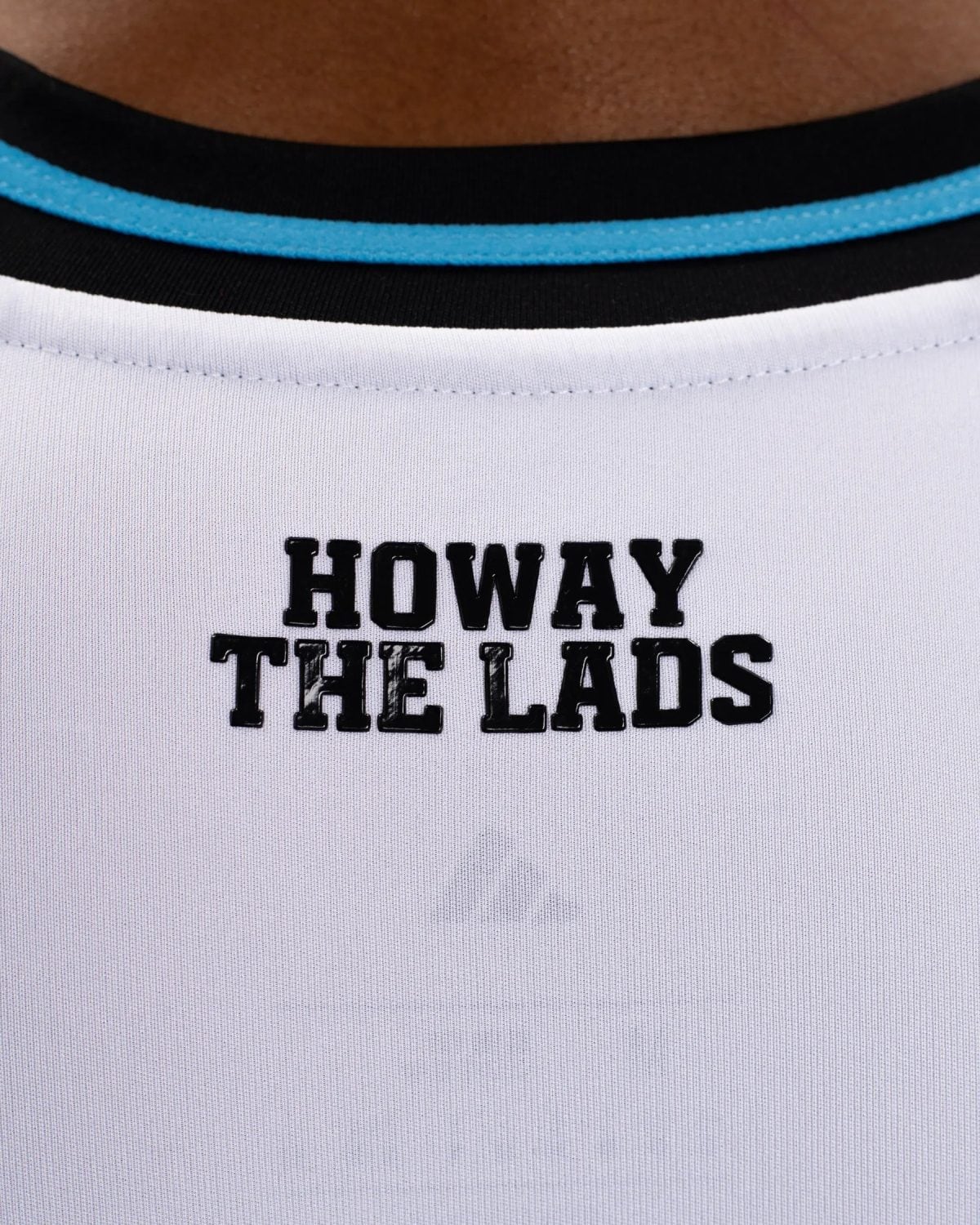 Close-up of a white T-shirt neck with a blue trim and black block letters reading “HOWAY” on the top line and “THE LADS” below.