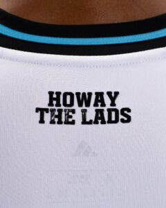 Close-up of a white T-shirt neck with a blue trim and black block letters reading “HOWAY” on the top line and “THE LADS” below.